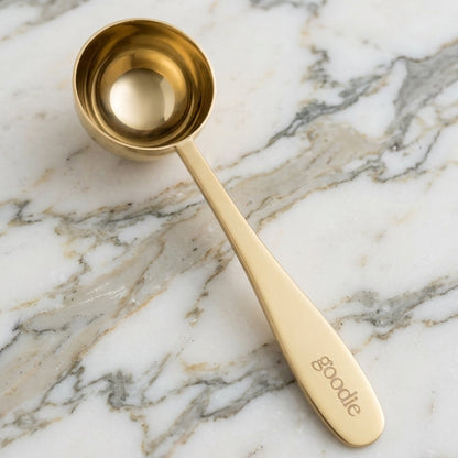 A gold-colored metal measuring spoon with a long handle engraved with the name 'Goodie', resting diagonally on a white and gray marble surface.