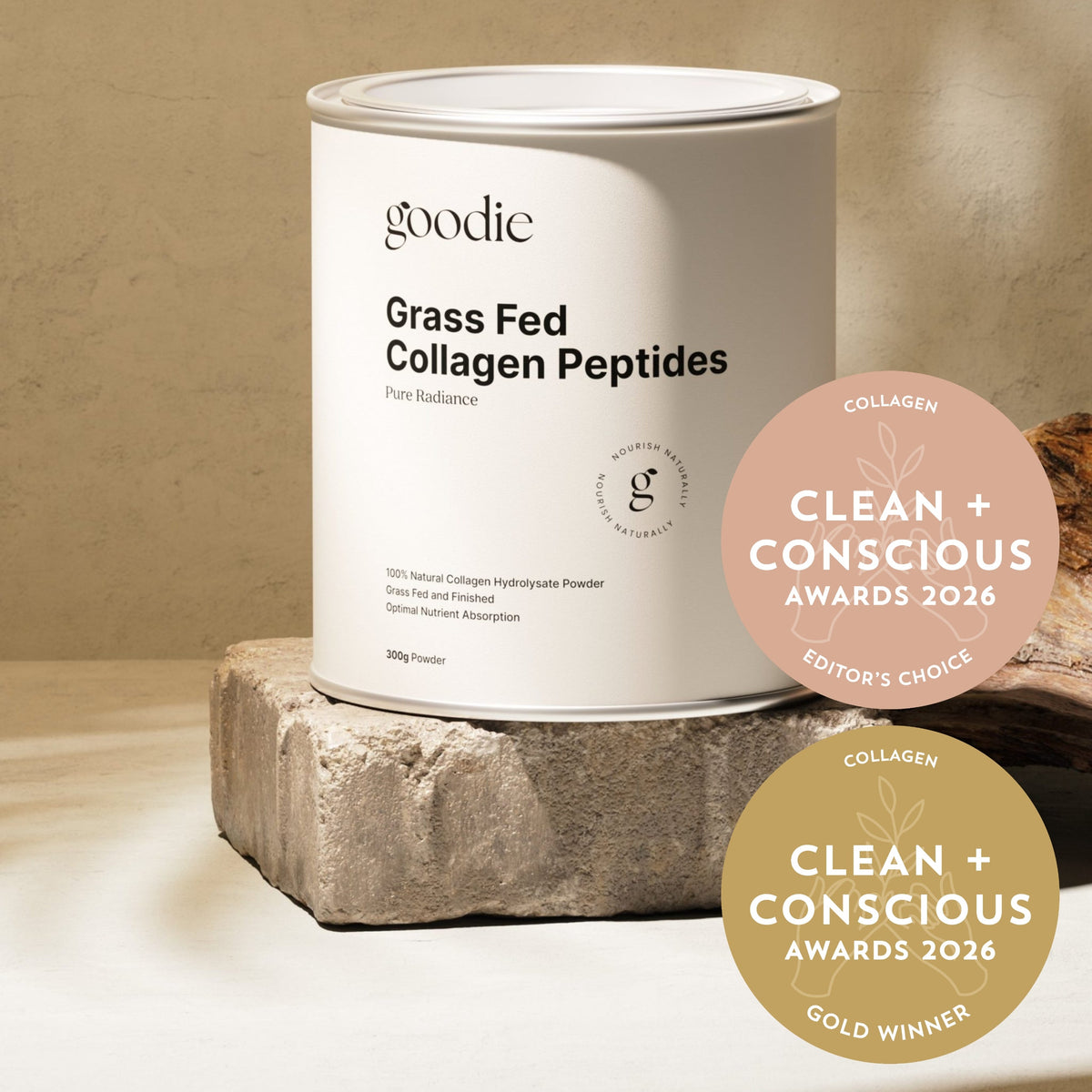 A white cylindrical canister of Goodie Grass Fed Collagen Peptides sits on a rough stone pedestal against a warm beige textured background. Two circular award badges for Clean + Conscious Awards 2026 are overlaid on the right side.