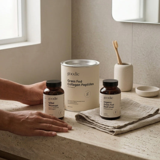 A person's hands rest on a beige stone countertop next to three Goodie brand supplement containers: a large canister of Grass Fed Collagen Peptides, a bottle of Vital Woman collagen, and a bottle of Organics Beef Liver. The scene is set in a modern, softly lit bathroom with a matching beige stone sink and a wooden toothbrush holder in the background.